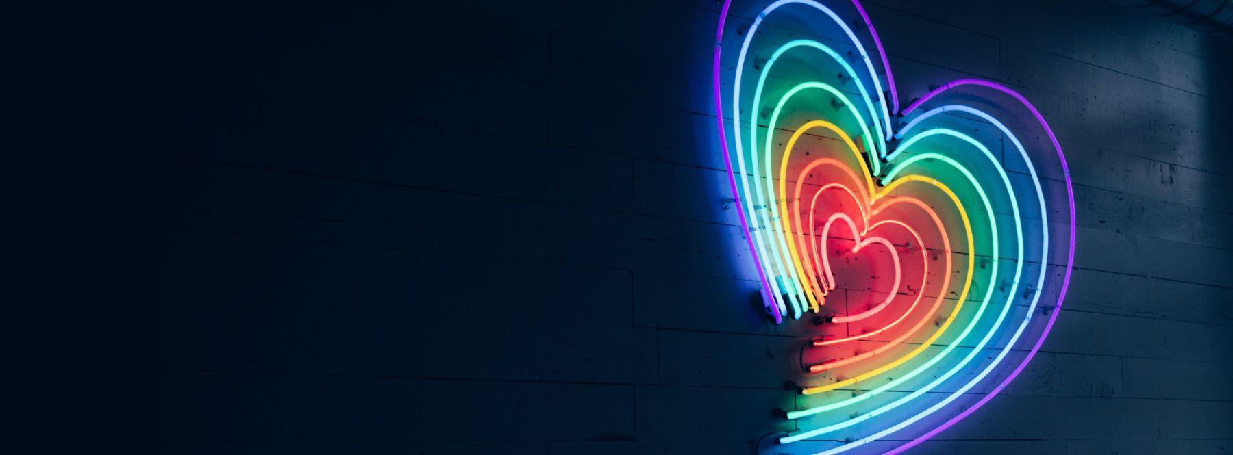 a dark interior space illuminated by a neon sign. The neon is shaped into 16 arcs, 8 on each side forming a heart. The arcs are in a rainbow gradient to symbolise support of the LGBTQIA+ community.