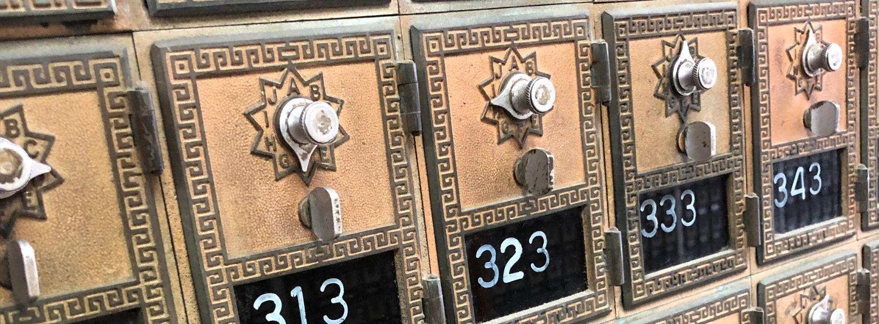 A close-up image of a bank of post office boxes from the Westminster, CO post office. The boxes are portrait-oriented rectangles. The bottom third of each box is a black field with the box number unevenly-printed in white, typewriter-style text. The top two-thirds of each box is a bronze color with a keyhole and latch. Each box is bordered in a greek-key-style pattern. Photo by Christopher Dowis, instagram @drmtch1. 